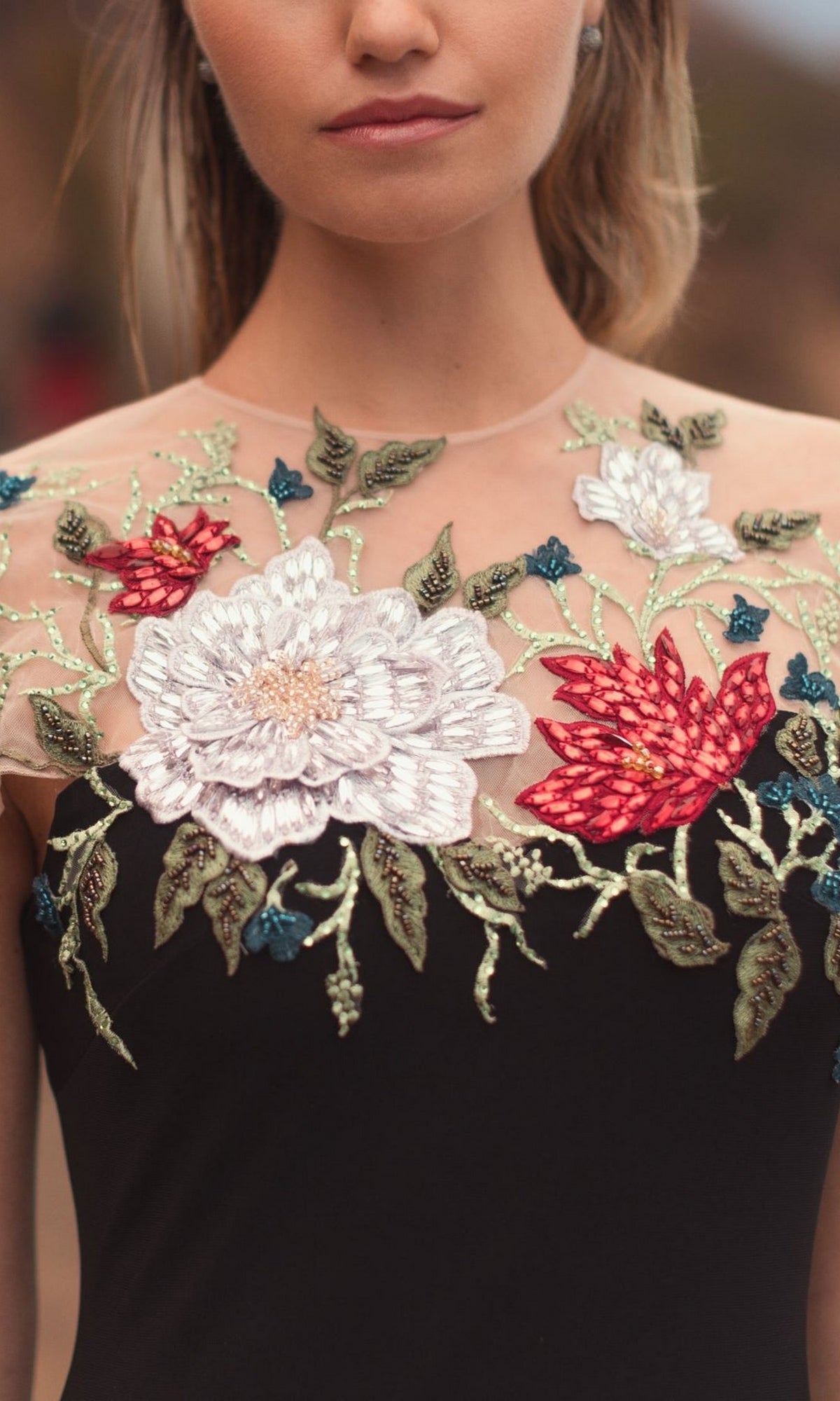 A woman wears the Floral Long Black Formal Dress: Mac Duggal 11817, featuring a sheer neckline and elegant embroidered white and red flowers with green leaves.